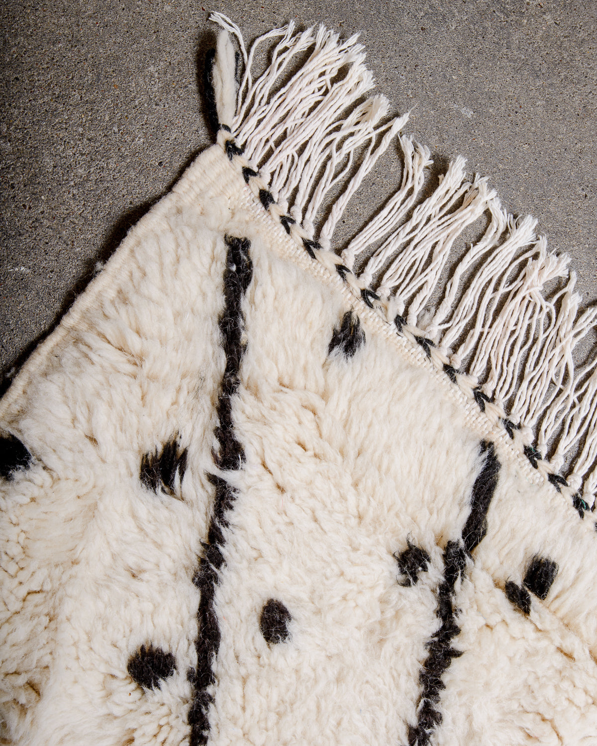 Corner of rug made of fluffy cream wool pile with thick black lines and large black polka dots and white cotton tassels.